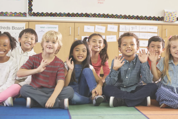 diverse group of children sitting in a classroom waving